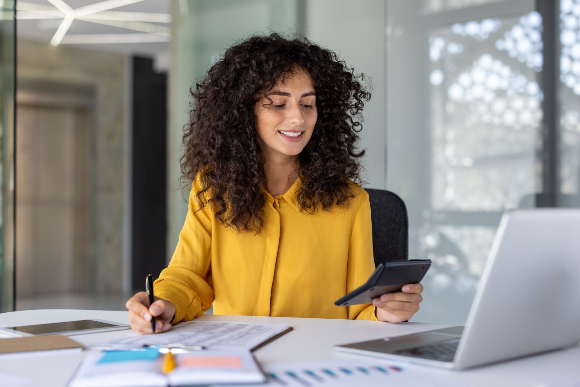 Confident Latin American businesswoman financial accountant using calculator and pen at modern office desk with laptop, showcasing multitasking and professionalism in corporate environment.